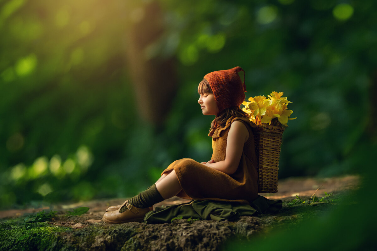 A girl sits on a moss-covered log with a basket full of daffodils in an enchanted forest portrait taken by Kara Reese Photography.