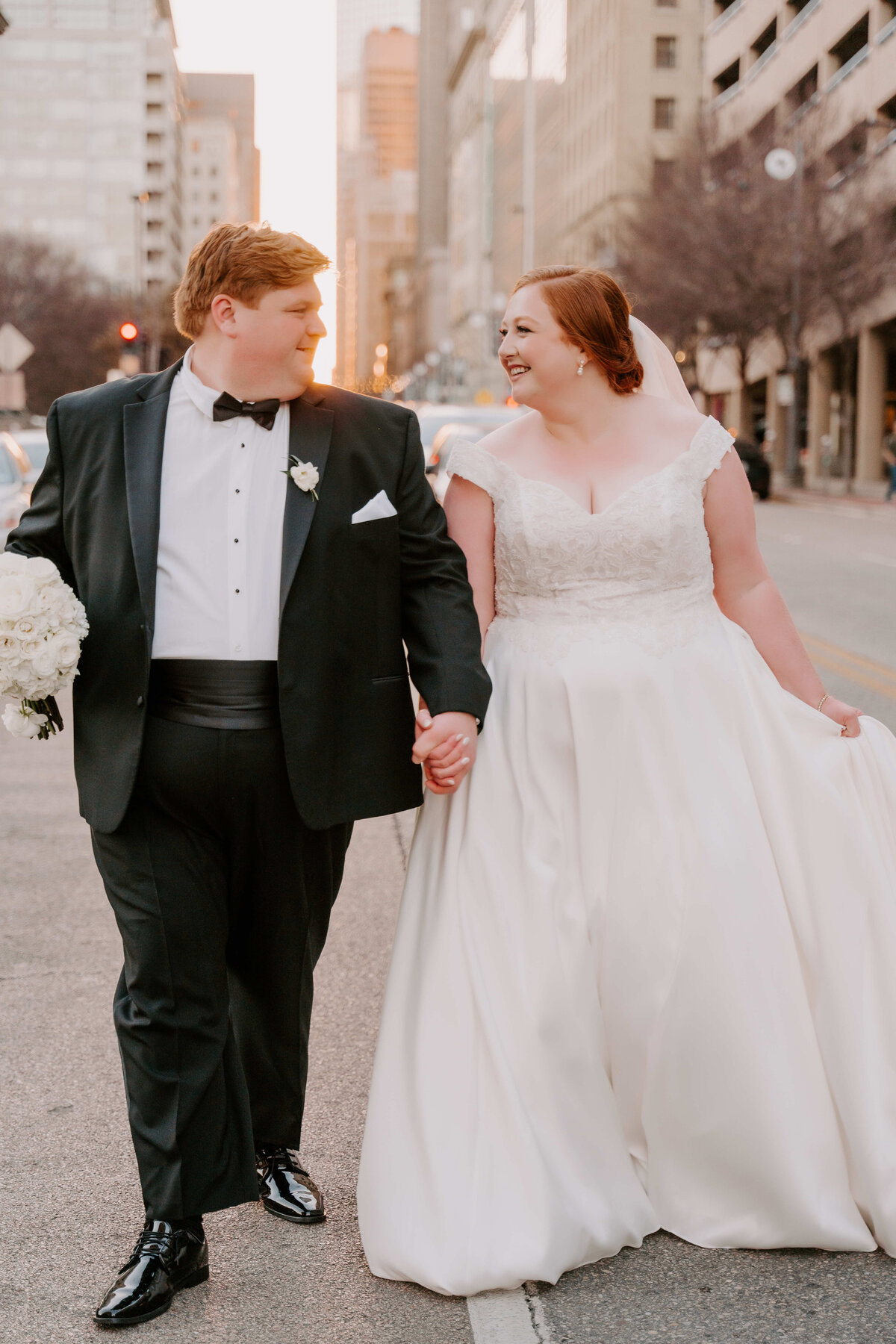 Bride and groom smiling and holding hands, walking down a city street at sunset. Bride in a white gown, groom in a black tuxedo. Romantic and joyful atmosphere.