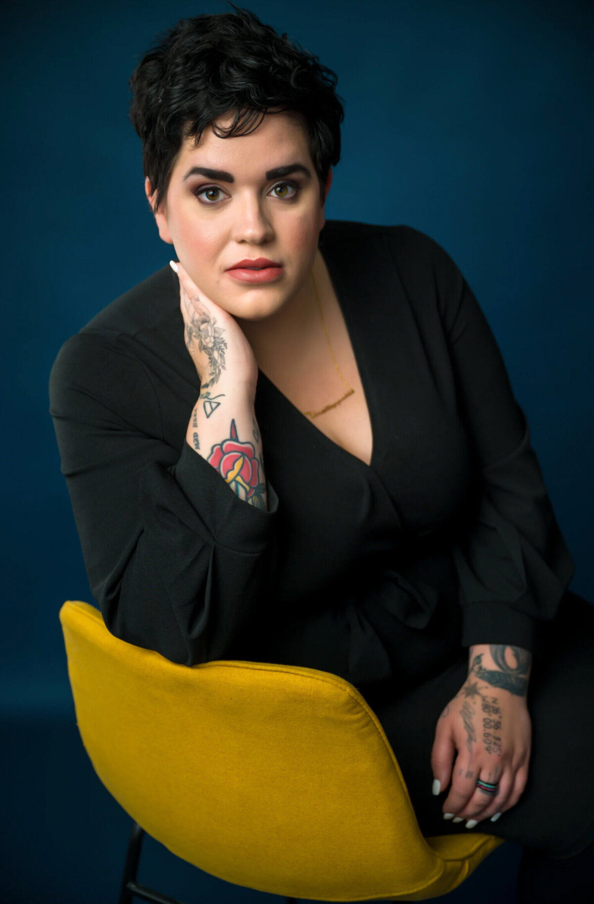 Woman wearing a black shirt sitting on a yellow chair on a blue backdrop, posing for a professional headshot