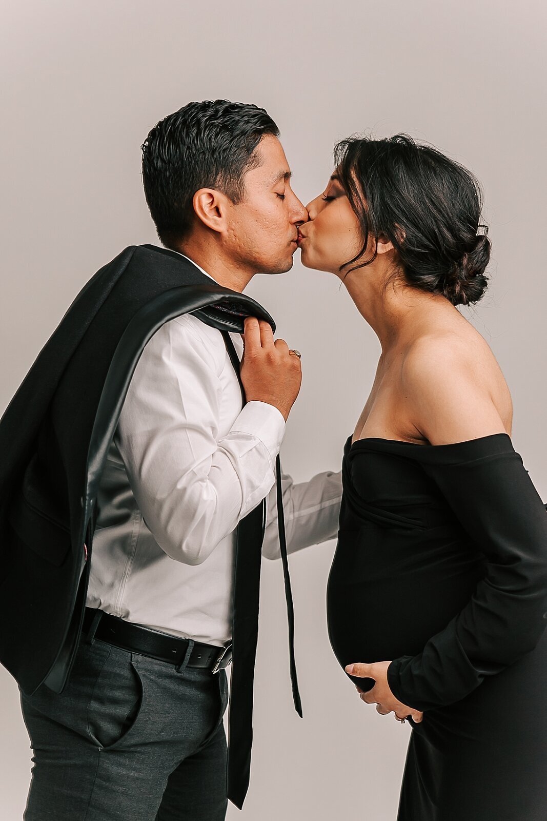 mom and dad kissing while she holds her belly. she is wearing a black dress and he is wearing a suit.