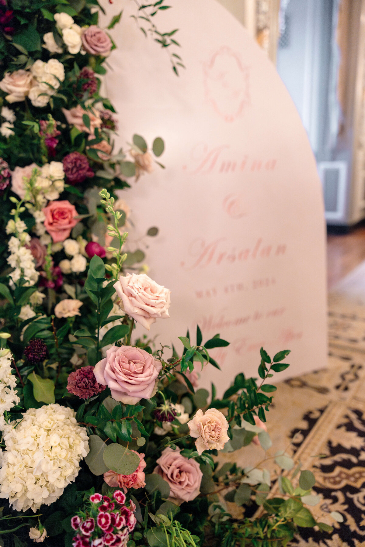A floral arrangement featuring pink, white, and burgundy flowers stands in front of a blurred sign with elegant cursive writing. The scene is set indoors with patterned carpet and soft lighting.