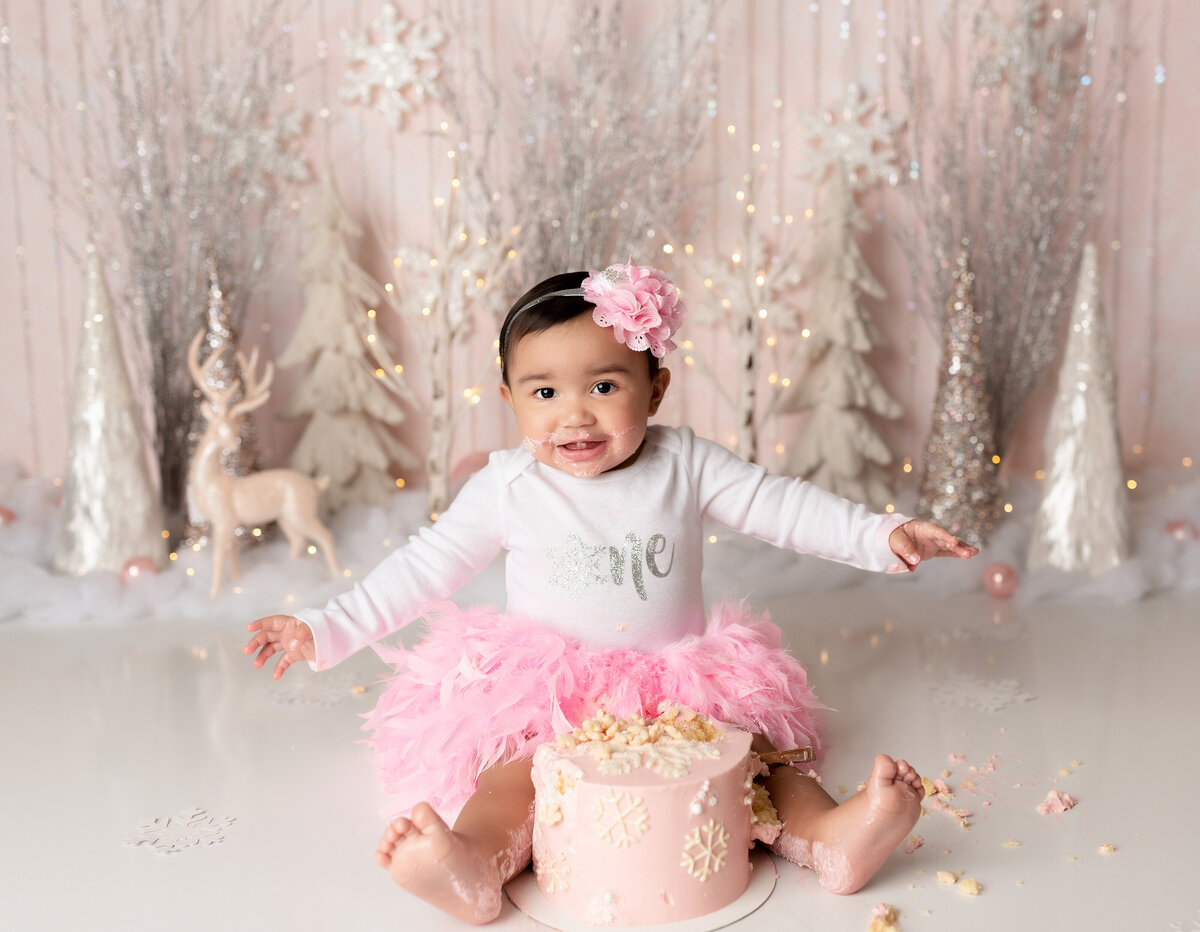 Winder Wonderland Cake Smash at West Palm Beach cake smash photography. Baby girl in a feather skirt is sitting behind the cake with her legs covered in icing, looking at the camera. The cake is pink with white snowflakes. In the background, there are white and silver trees, deer, and faux snow.