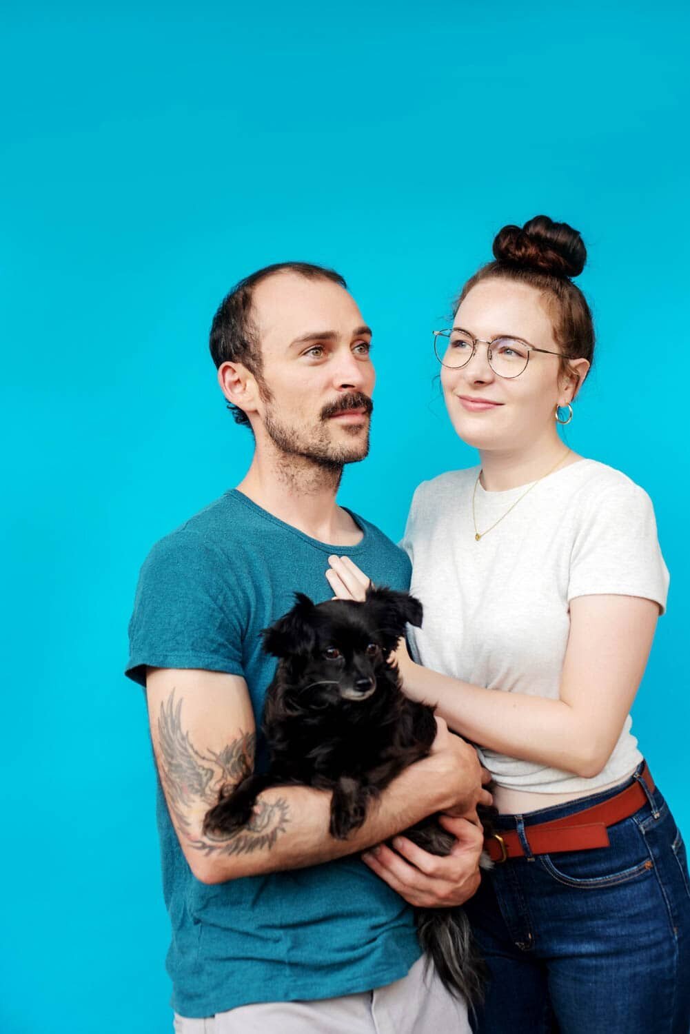 a man and woman holding a dog standing in front of a colorful bright blue background look in opposite directions