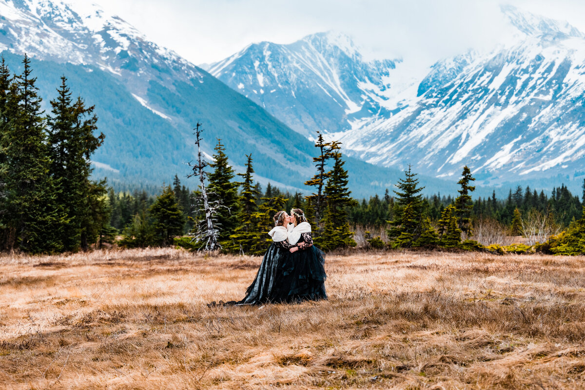 Tow brides, both wearing black wedding gowns, white fur stoles, and flower crowns, kiss in a meadow following their Alaska elopement ceremony during bridal portraits.