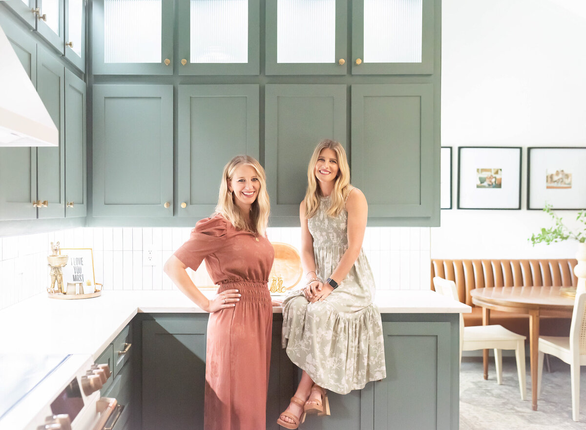 owners of an interior design company in a kitchen they designed with a pretty blue-green cabinet color and one sister is sitting on the counter and the other one is standing next to her