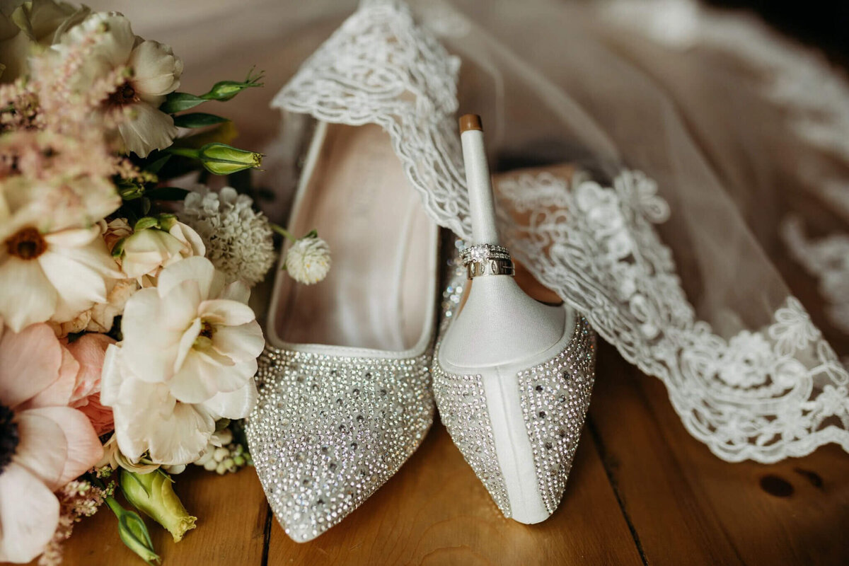 Photo of sparkly white heels with wedding rings on them laying with a veil and pink florals