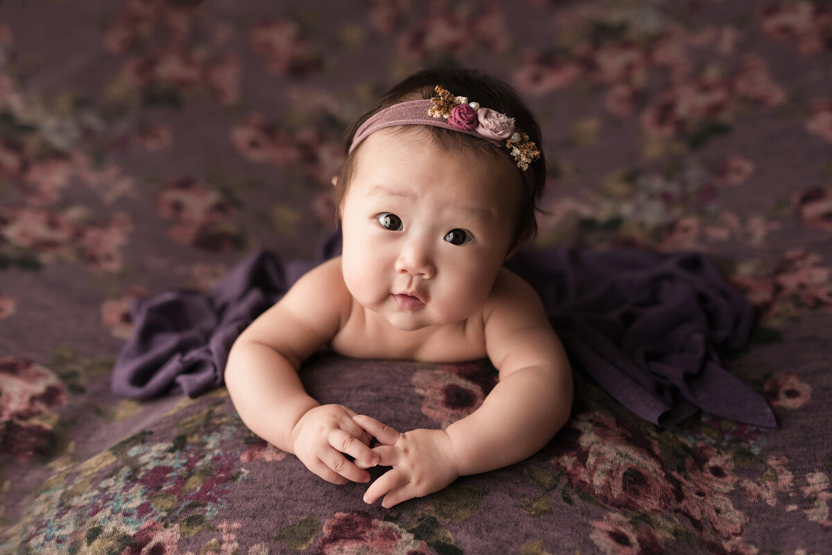 3-month milestone photoshoot at top West Palm Beach and Jupiter baby photographer. Asian baby girl is laying on her belly with her arms in front of her and looking up at the camera. Baby is laying on purple floral fabric .