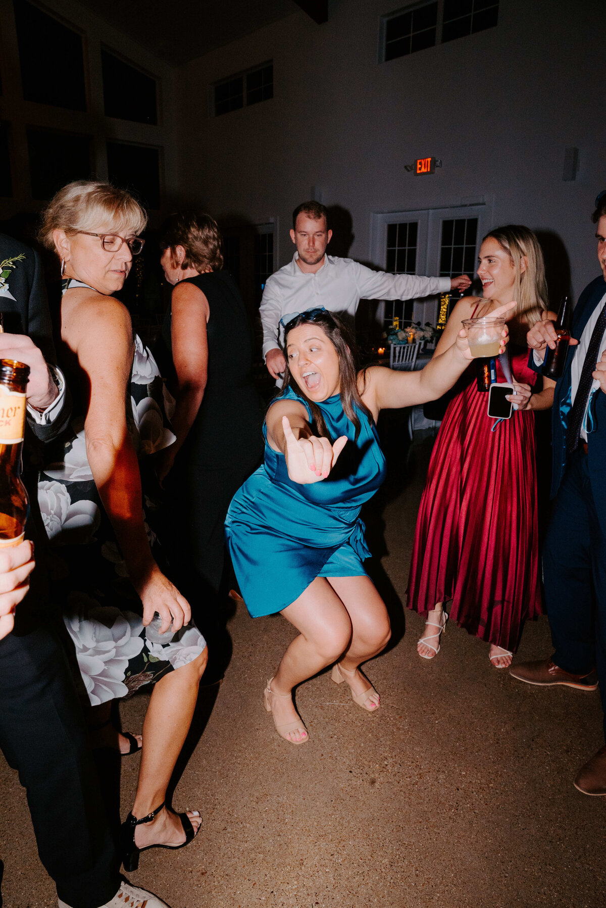 A lively dance floor scene at a party, featuring people in formal attire. A woman in a blue dress energetically poses, surrounded by smiling guests.