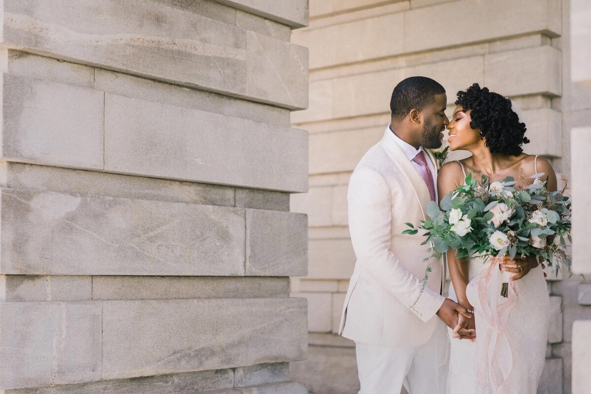 A bride and groom stand close, sharing a tender moment against a classical architectural backdrop