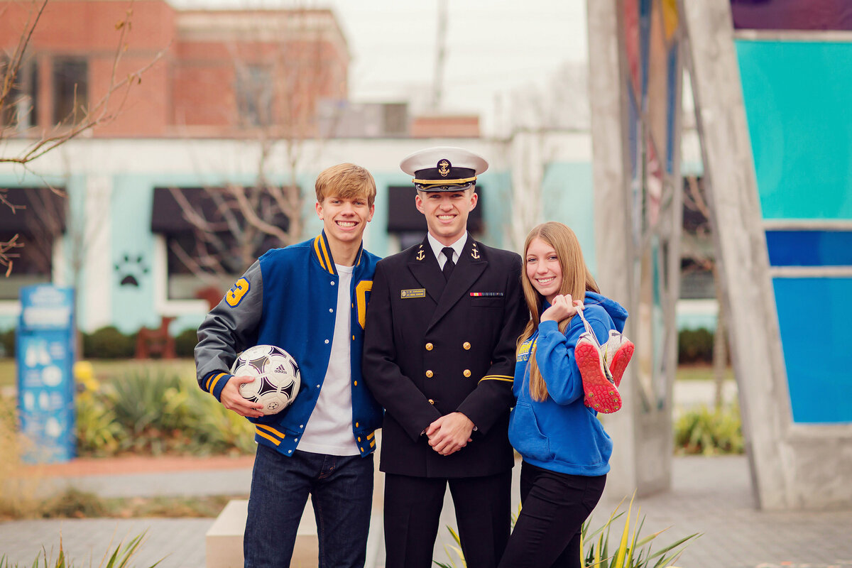Sibling in their gear in Carmel. One is wearing a letterjacket holding a soccer ball. One is in his Navy uniform. The other is holding her track shoes.