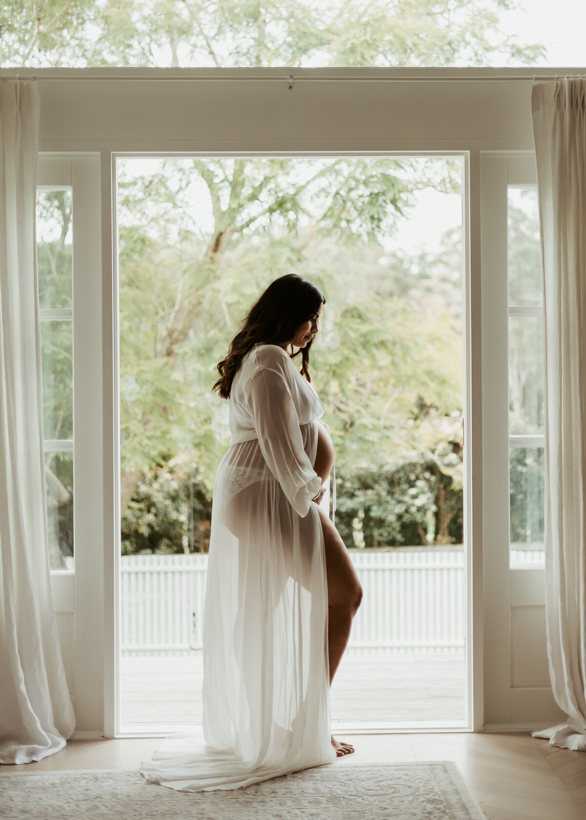 Pregnant woman in see-through white robe posing in doorway of opened French white doors