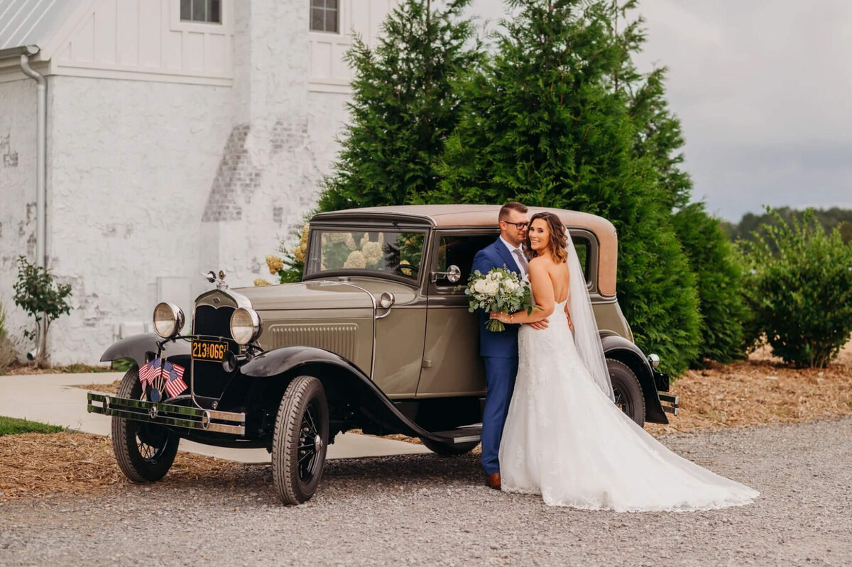 Photo of a bride and groom hugging while leaning up against a vintage car with American flags on the front