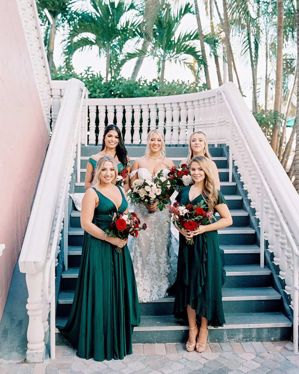 Bridal party standing on the stairs of the Don Cesar Hotel in St. Pete Beach, FL, during a December wedding. The bridesmaids are in emerald green floor-length satin gowns, holding red and white floral bouquets.