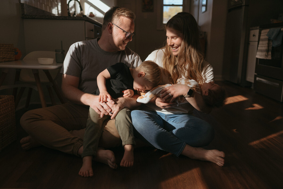 A mom holding a newborn baby,  a dad and a toddler boy  sit in bright sunlight snuggling and laughing