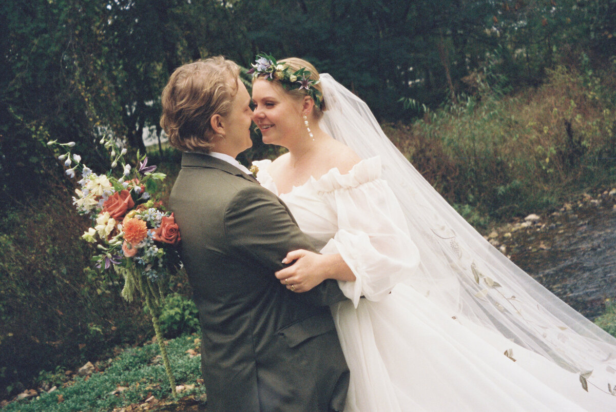 Bride and groom embracing beside a wooded creek at an Arkansas wedding, featuring a flowing off-the-shoulder gown and a vibrant garden-style bridal bouquet with dahlias, roses, ranunculus, and whimsical greenery designed by a Northwest Arkansas florist.