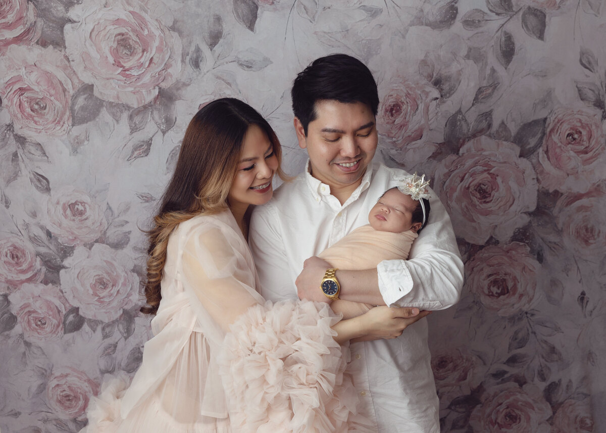 A happy mom and dad smile down at their sleeping newborn daughter in dad's arms while standing ina. studio against a floral backdrop