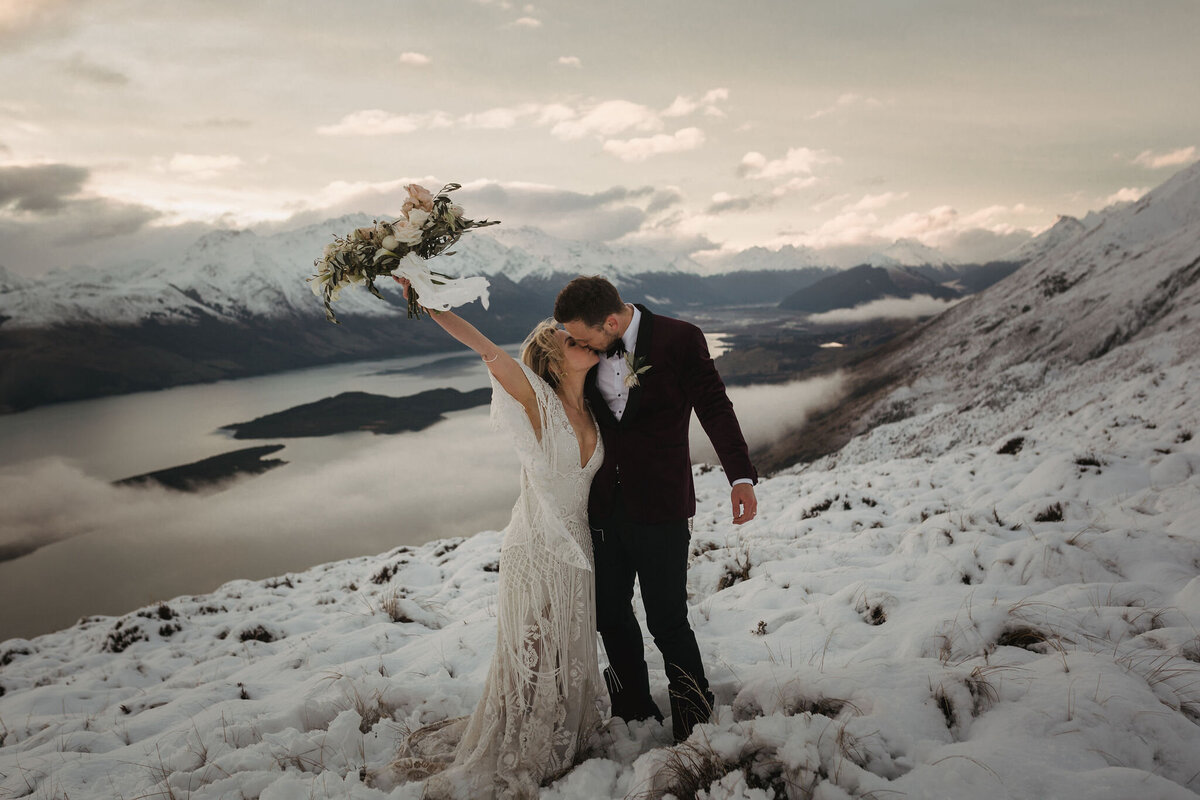 The Vase Floral Co - bride and groom kiss on top of Queenstown mountain - bride holds bouquet in the air