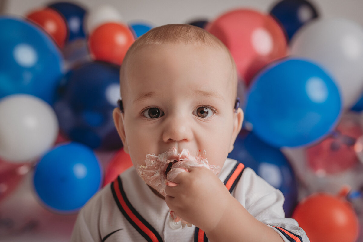 One year old baby boy in Atlanta Braves jersey eating a birthday smash cake, closeup shot of him with red, white and blue balloon garland in background