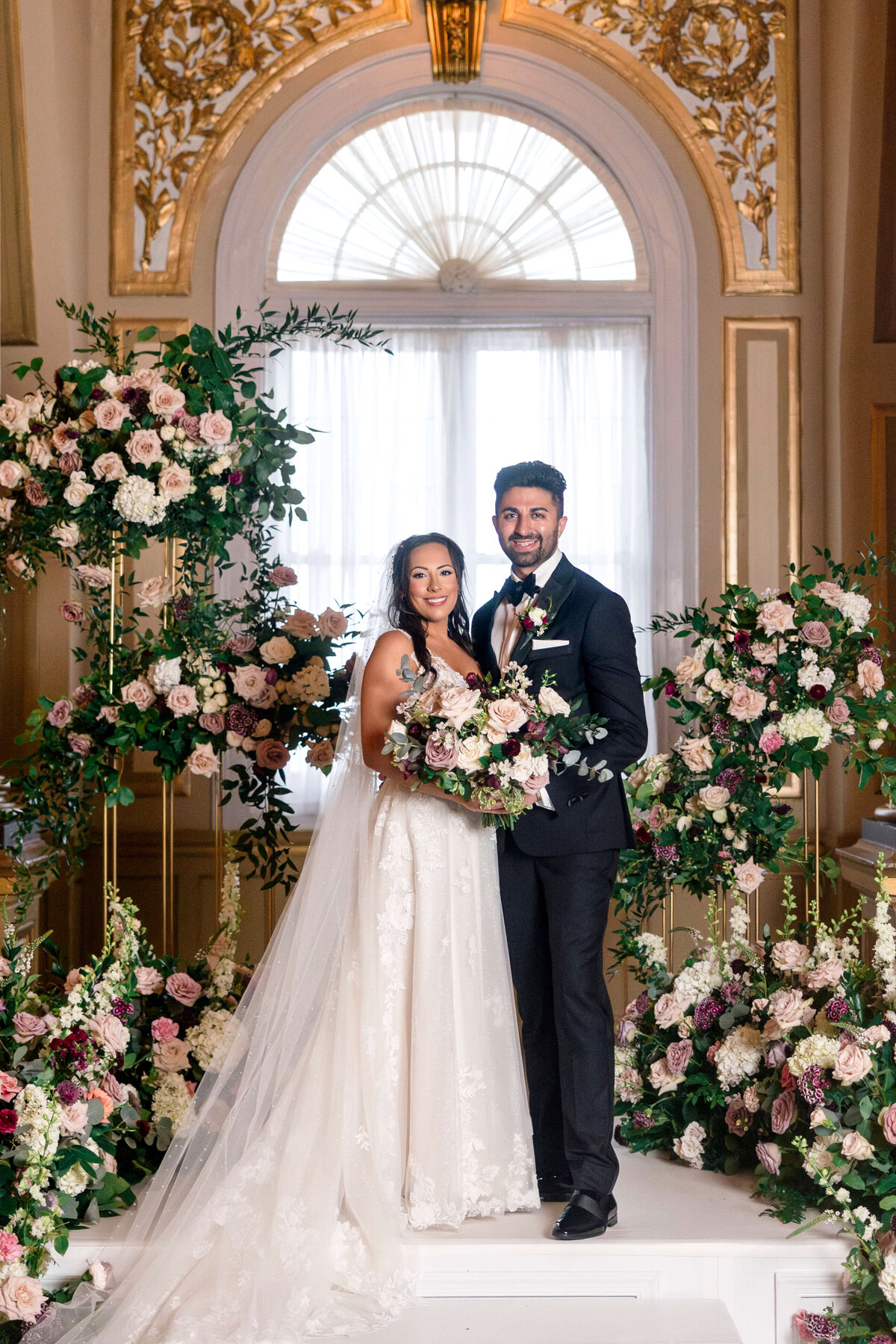 A bride and groom stand together on a decorated stage with an elegant floral backdrop. The bride wears a white gown and veil, holding a bouquet, while the groom is in a black tuxedo. They are smiling in front of a large arched window with ornate details.