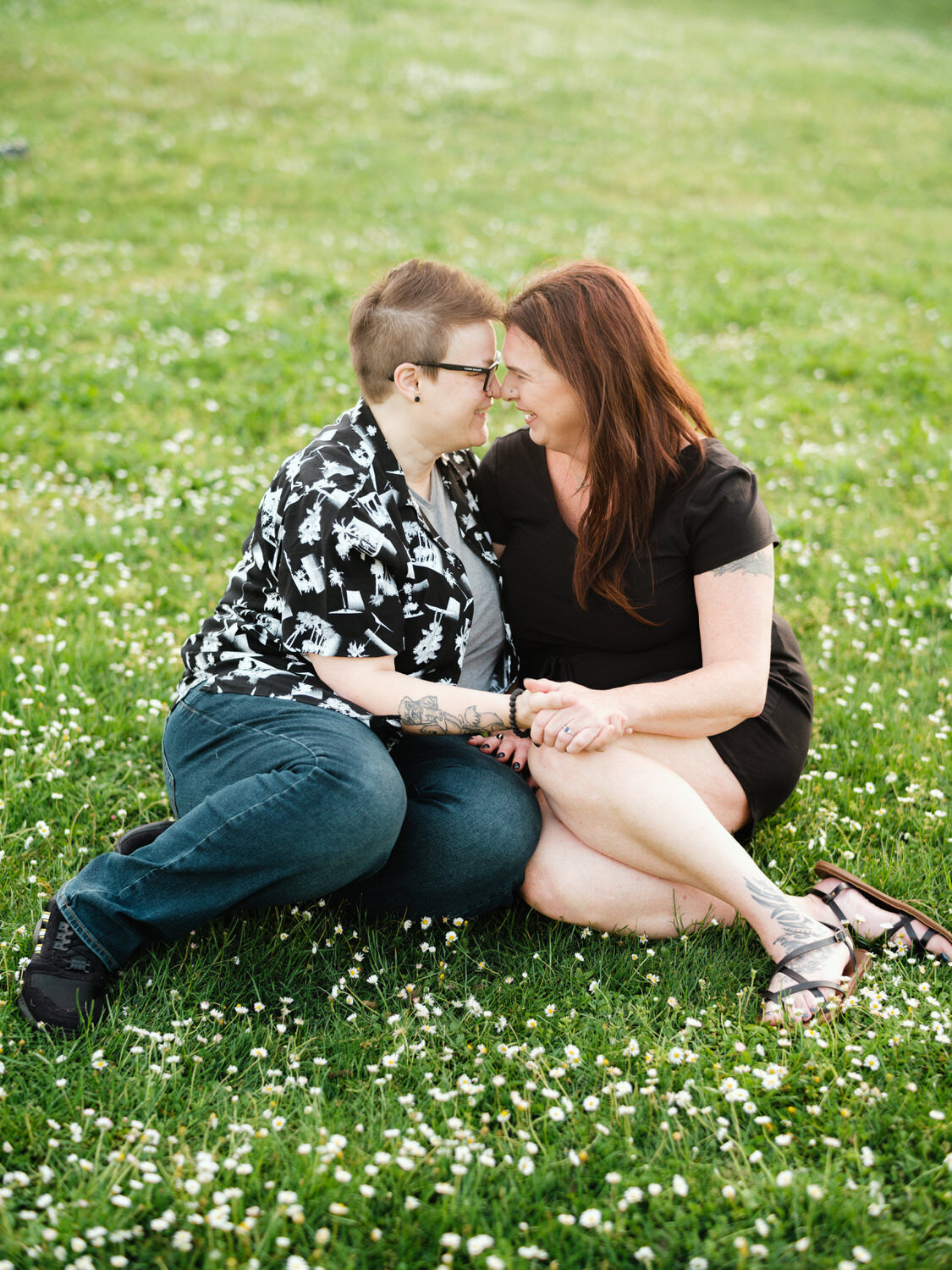 LGBTQ couple smiles while sitting in the grass during an engagement photo session