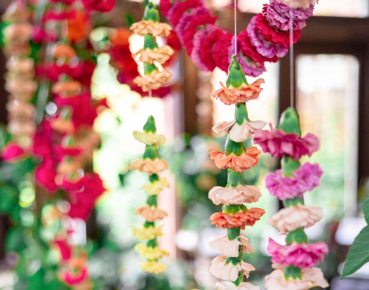 Colorful floral garlands hang in a display, featuring orange, yellow, pink, and red flowers. The garlands are set against a blurred background with greenery, creating a vibrant and festive atmosphere.