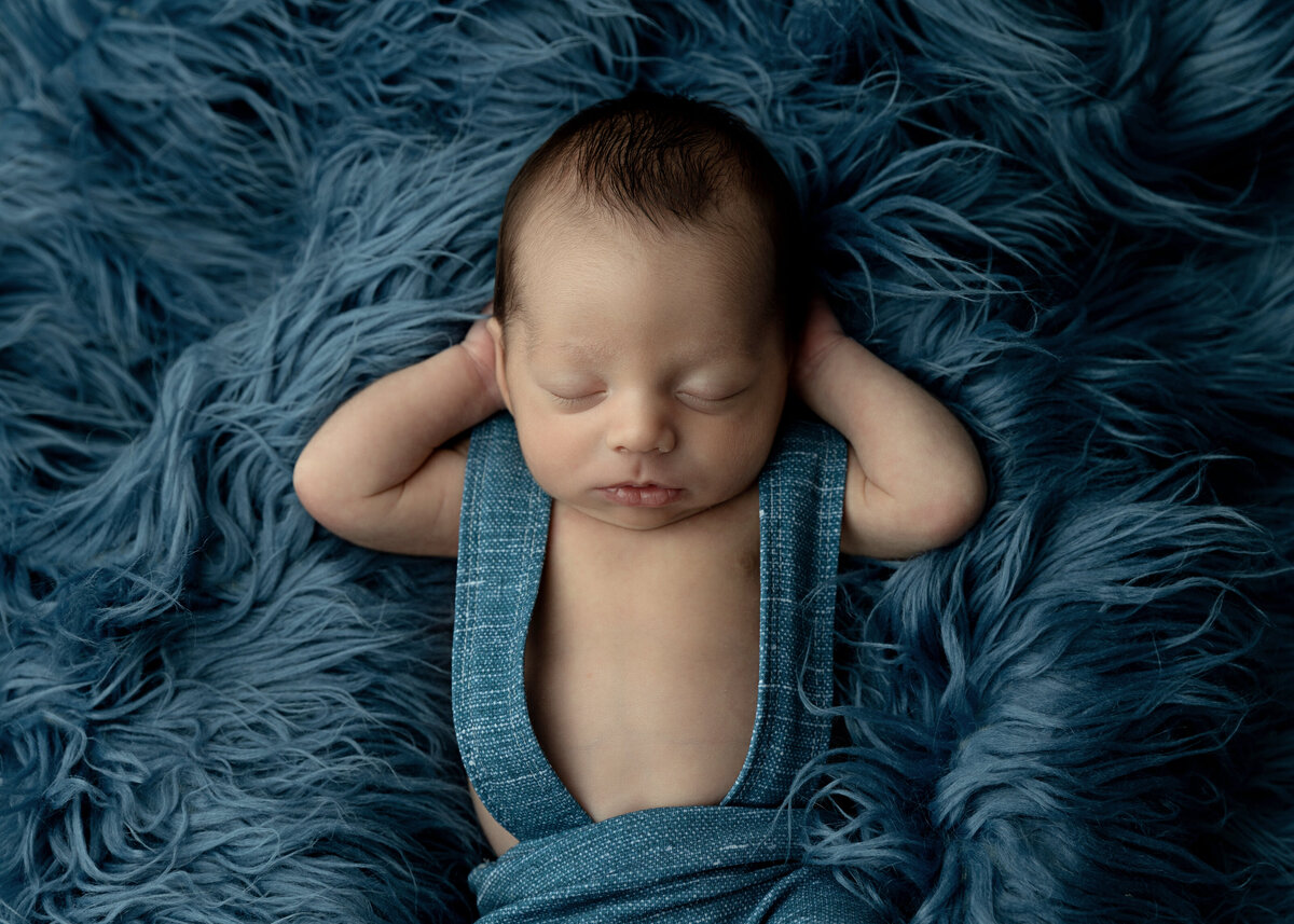 A newborn baby sleeps with hands behind his head in blue overalls during a NJ Newborn Photography session