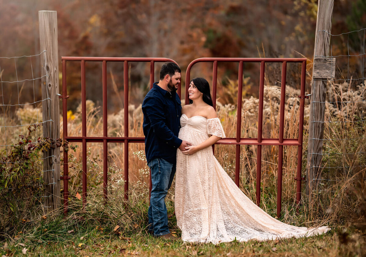An Adorable expecting couple gaze into each others eyes while standing in front a red gate in the country outside of Asheville