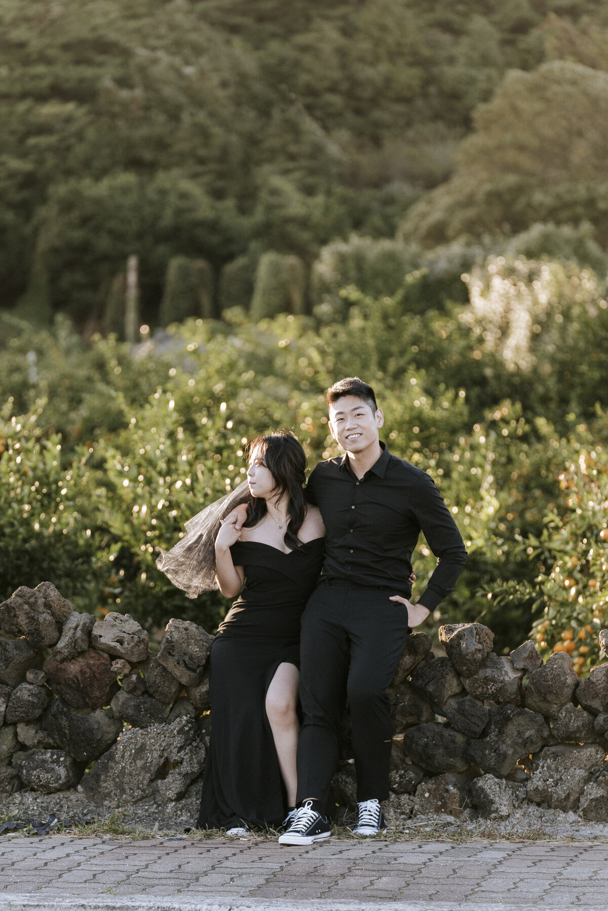the bride wears black dress with slit and a little black veil then the groom wears an all black outfit while sitting on the pile of rocks
