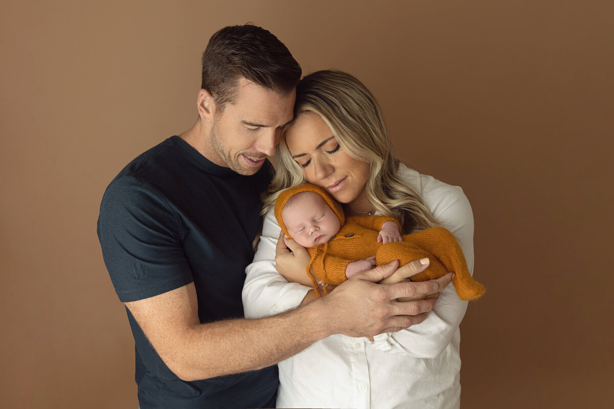Happy new parent snuggle with their sleeping newborn baby in an orange onesie while standing in a NJ Newborn Photography studio