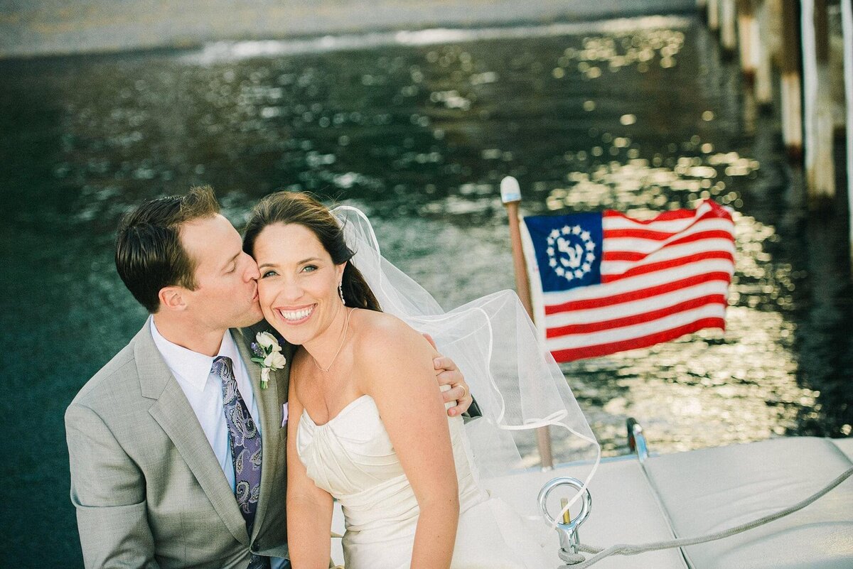 newlyweds sitting on boat with groom kissing his bride with usa flag behind in Lake Tahoe