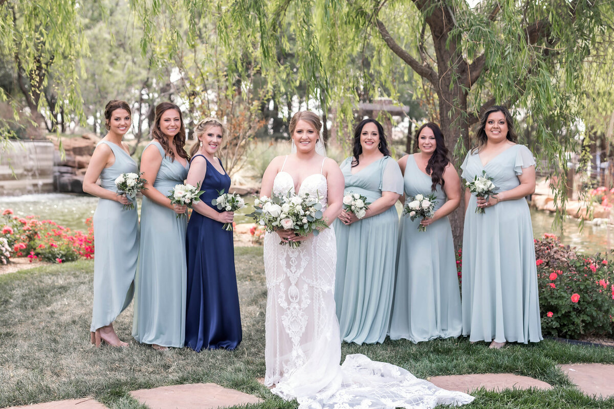 Bridal party all looking at the camera and holding their bouquets. Bride is wearing a strapless lace dress and the bridal party is wearing seafoam green and a navy satin dress.