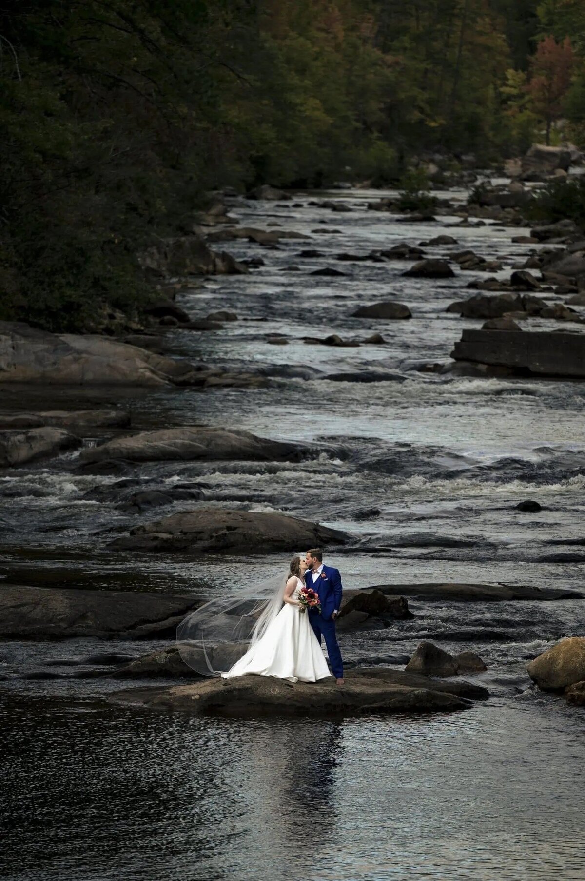 A bride and groom stand on a rock in the middle of a gently flowing river, surrounded by the tranquility of nature, encapsulating a moment of peaceful solitude.