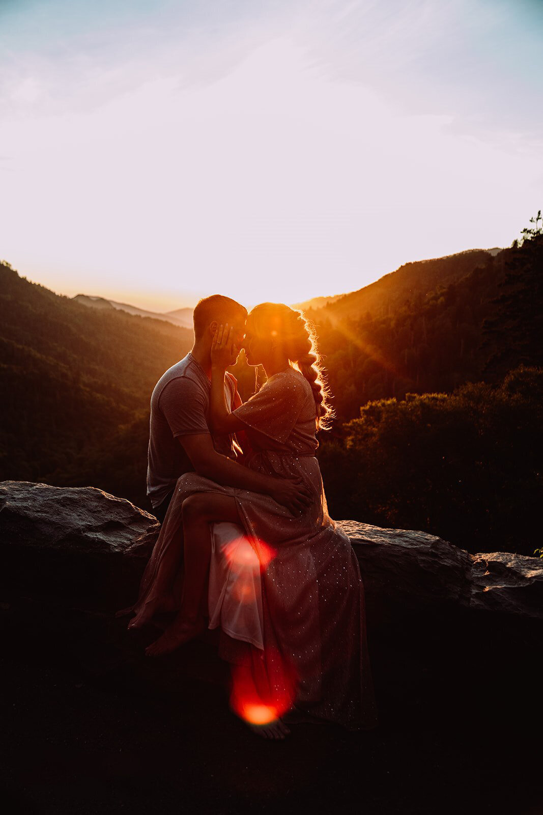 couple sitting on a wall looking at each other with the sun light and mountains behind them