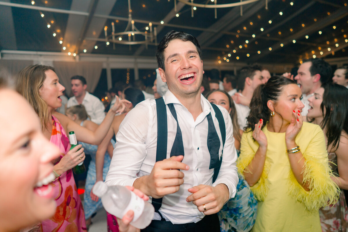 Lowndes Grove groom laughs on the dance floor with string lights and wedding guest in bright yellow dress with feather sleeves. Kailee DiMeglio Photography.