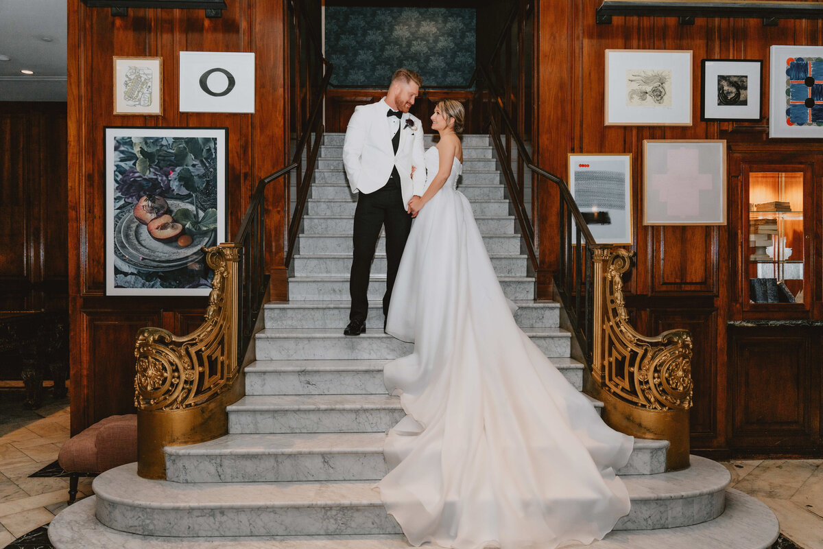 A bride and groom stand on an elegant staircase in a lavish setting. The bride wears a flowing white gown, and they look at each other lovingly.