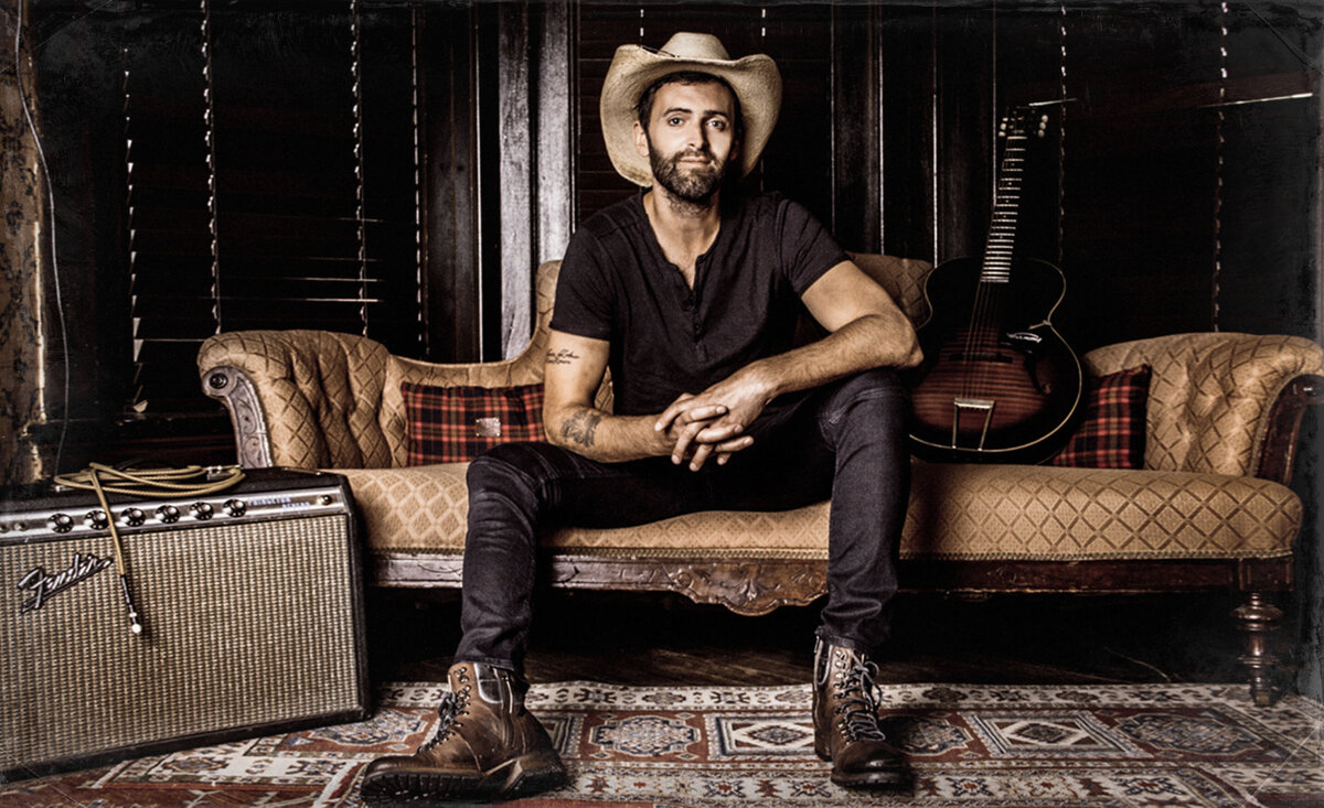 Male musician portrait Dean Brody wearing black outfit straw cowboy hat while sitting against gold sofa beside amplifier with guitar