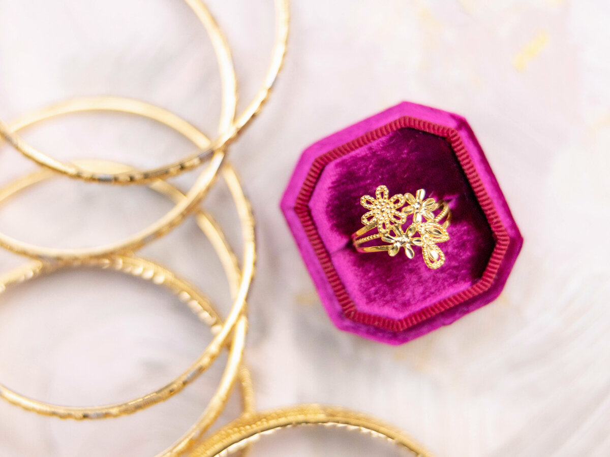 A gold floral ring in a purple velvet octagonal box, surrounded by several gold bangles on a light, textured background.