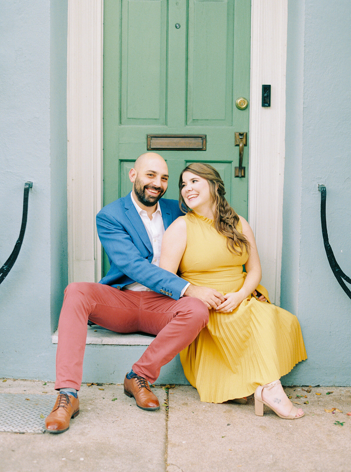 Downtown Chalreston colorful engagement session. Guy in burnt red pants and blue blazer. Girl in yellow dress in front of green door. Charleston film photographer.