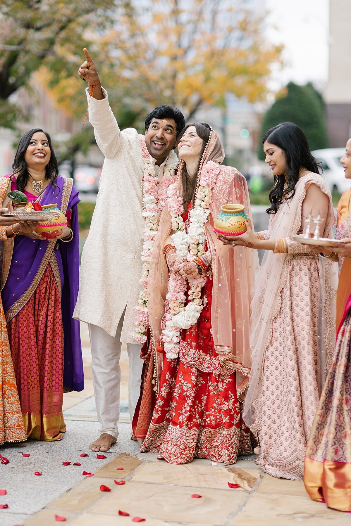 Indian groom wearing a white sherwani and blush floral varmala points to the sky as an Indian bride wearing a red and gold saree wearing a blush floral varmala looks up. Indian wedding guests holding offerings to the gods surround them at there Hindu wedding in Nashville, TN