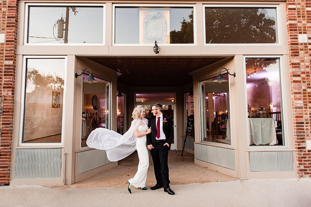 The bride and groom stand in front of a storefront with many glass windows in an exposed brick building. The groom is wearing a black suit with a white shirt and dark red tie with his hands in his pockets. The bride has her arm interlocked with his as she leans in. She is wearing a sheath  floor length gown with a cape that is blowing in the wind behind her.