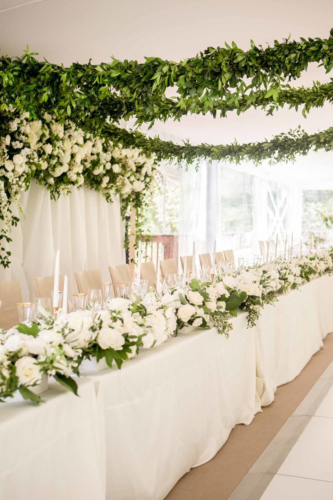 Grand tent wedding head table with white floral  and green garlands on ceiling