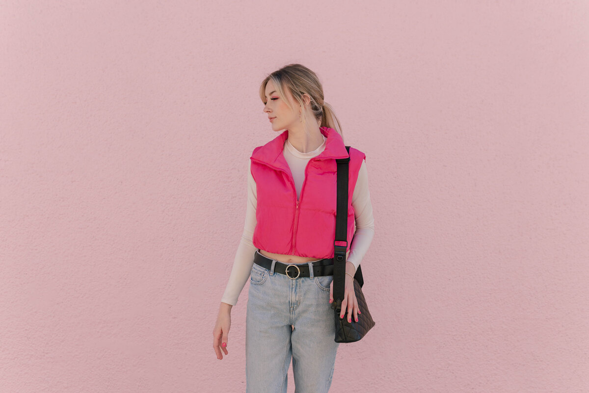Sporty Barbie looks down over her shoulder while holding a black purse in front of a pink wall in the Montrose area