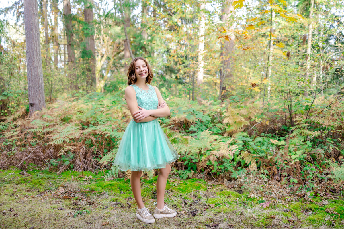 A teenage girl in a green dress proudly smiles while standing in a forest park trail