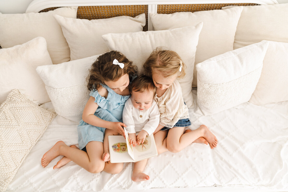 Three young siblings enjoying a storybook together on a cozy bed filled with cushions.
