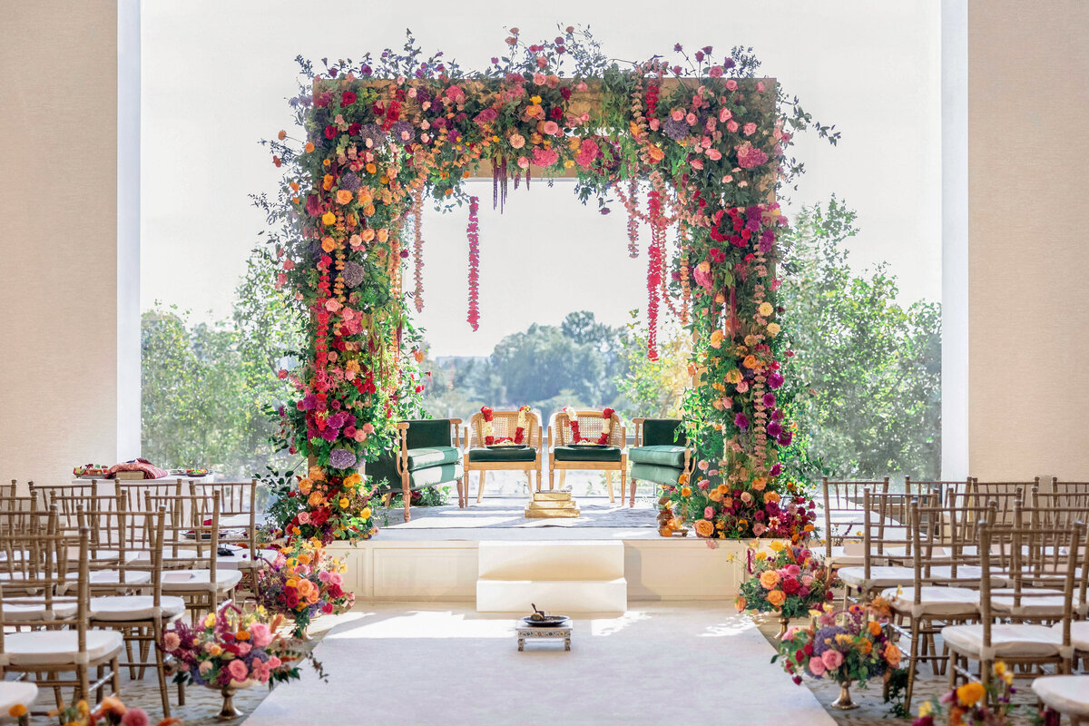 A beautifully decorated wedding mandap with an arch adorned with colorful flowers, including pink, orange, and purple blooms. Two green velvet chairs are set underneath. Rows of chairs line the aisle, and large windows reveal greenery outside.