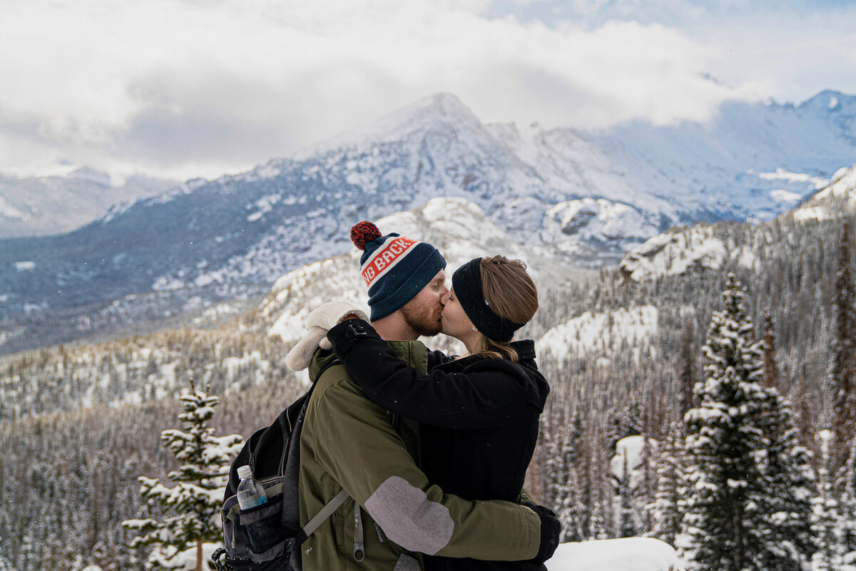 Man and woman in hats and jackets kiss in snowy Rocky Mountains.