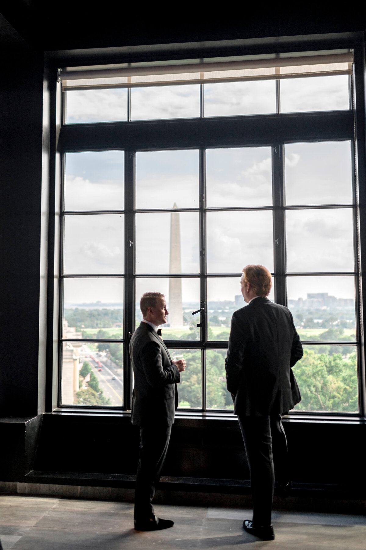 Two people in suits stand near a large window overlooking the Washington Monument. The window panes frame the view of the monument and a green landscape. The room is dimly lit, contrasting with the bright scene outside.