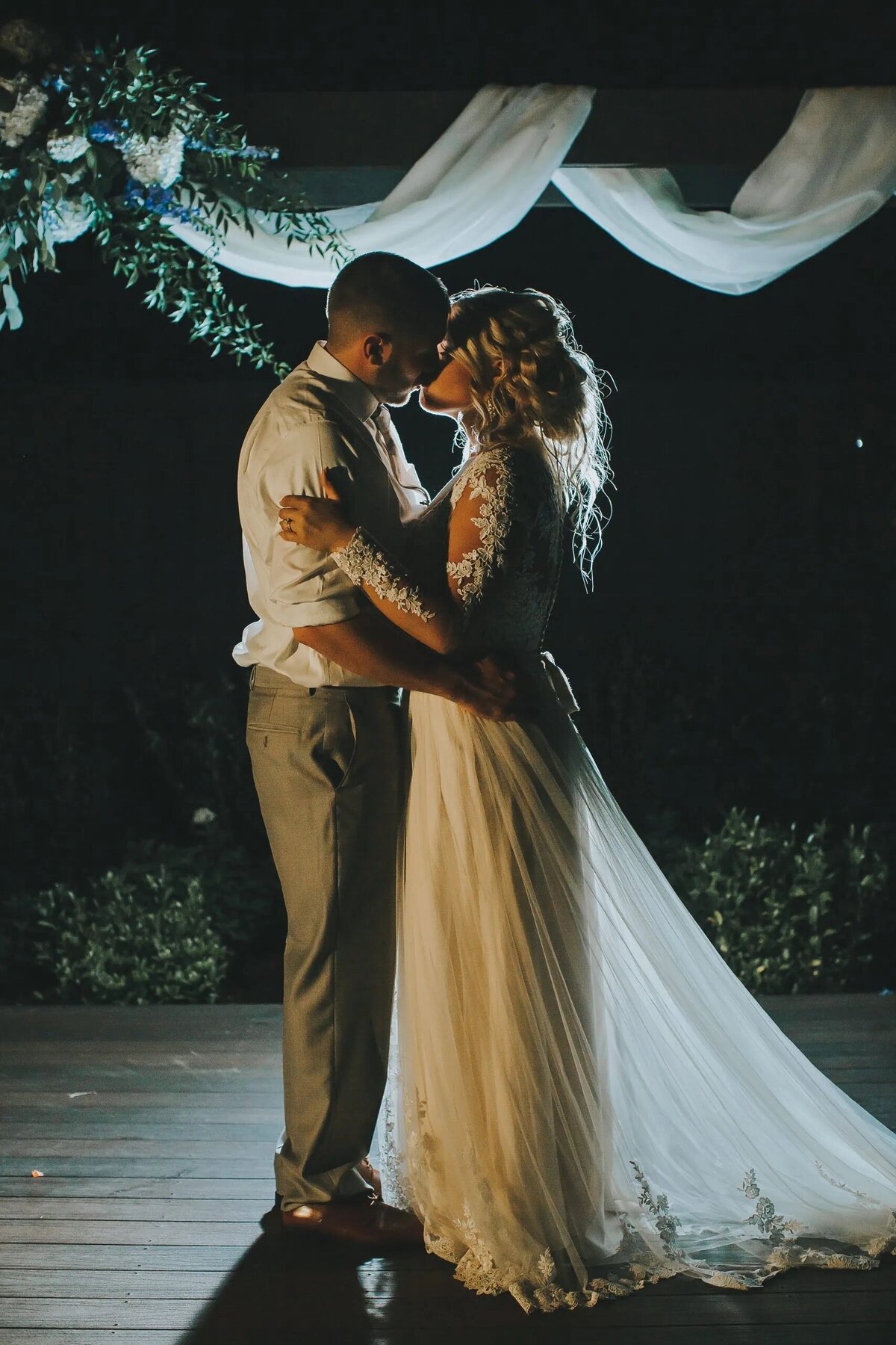 A bride and groom share an intimate dance under soft ambient lighting, with draped fabric above, in a romantic evening setting.