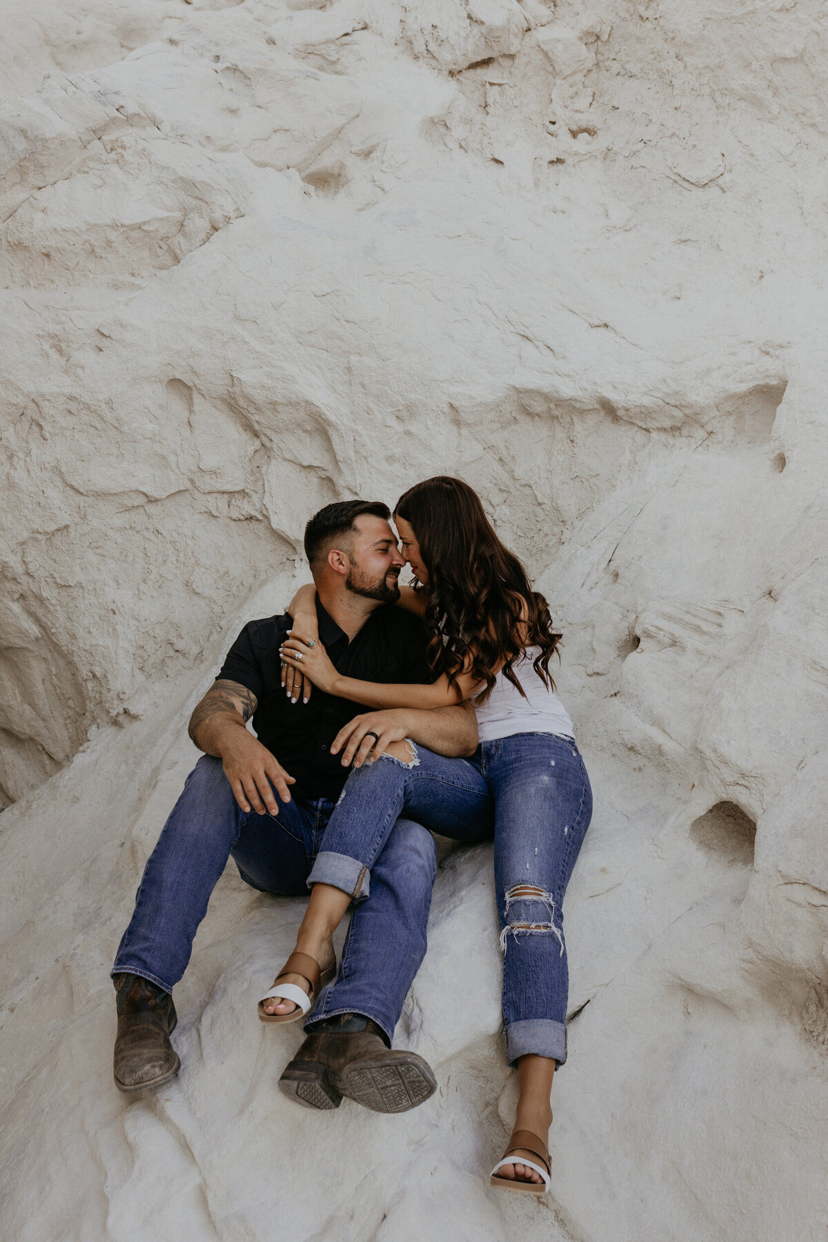 engaged couple sitting on a white rock together about to kiss