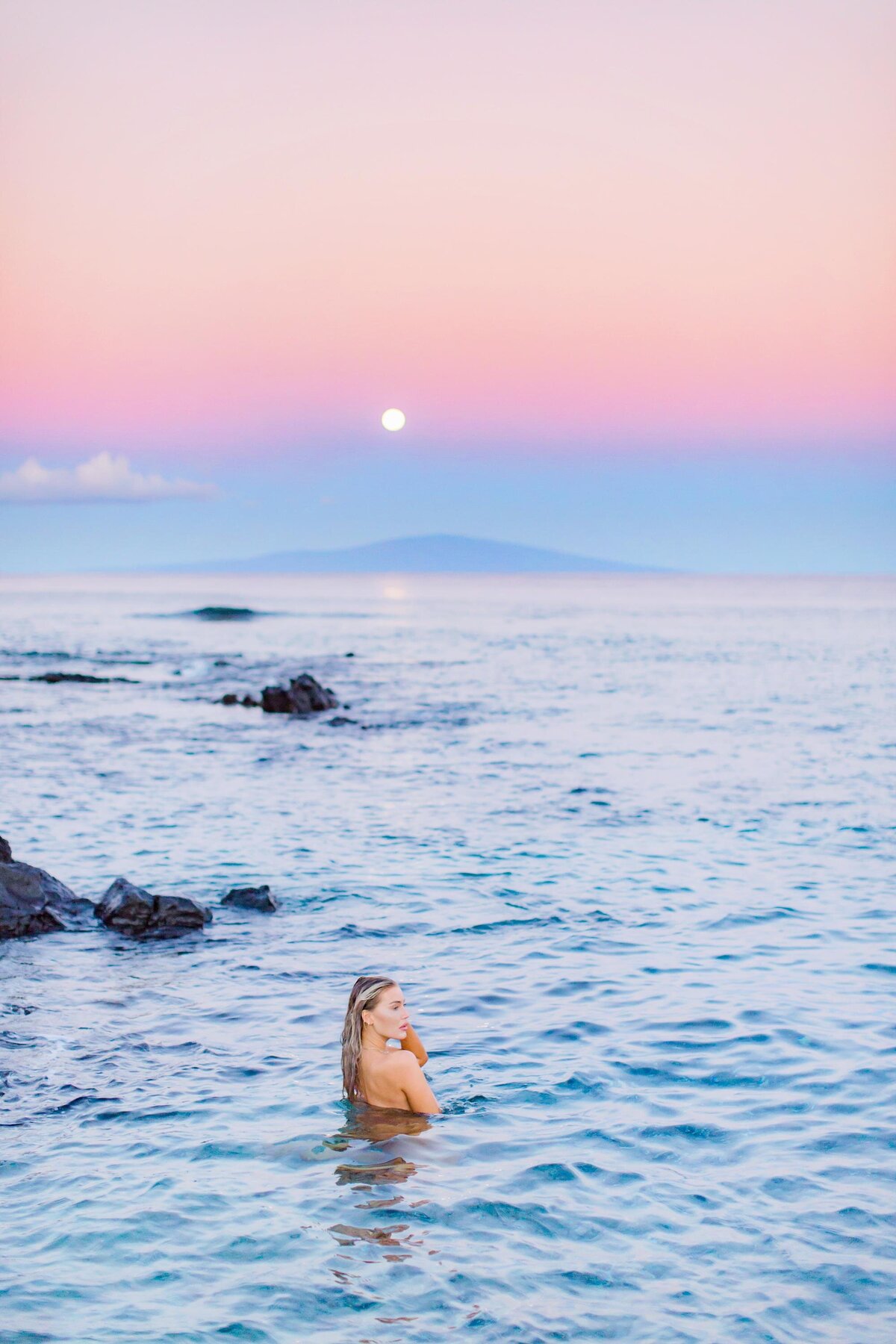 Blonde woman stands in the water and stares out at the ocean under a pastel sky